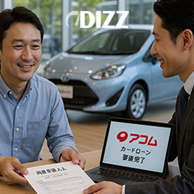 自動車のショールーム内で、カジュアルな服装の男性顧客が、スーツ姿の男性担当者と向かい合って座っています。両者とも笑顔で、担当者は書類を提示しています。テーブル上のタブレットには、**アコム（Acom）のロゴと「カードローン 審査完了」**という文字が表示されており、カードローンの審査が完了したことを示唆しています。